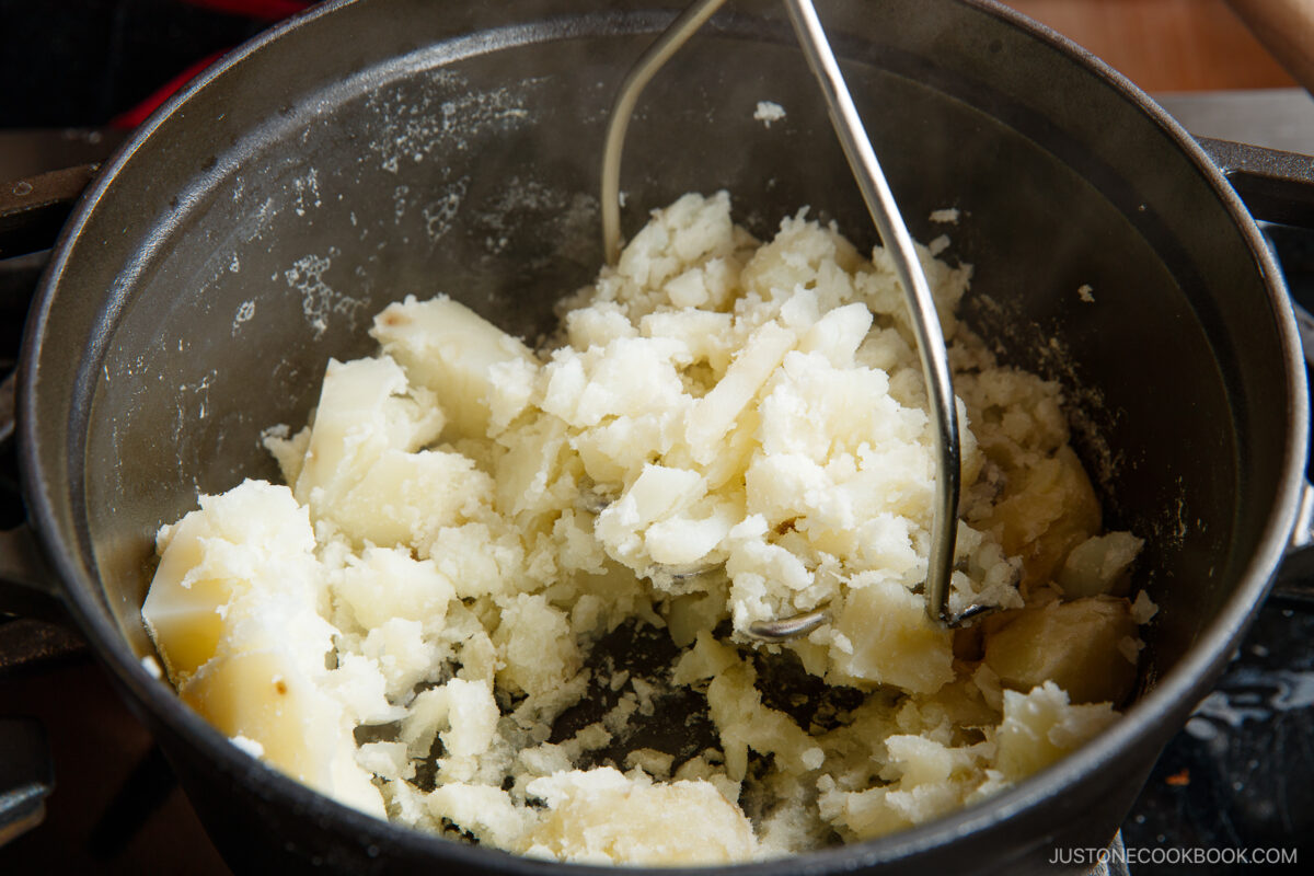 A close-up of a pot filled with cooked potatoes being mashed with a metal potato masher—fresh, steaming potatoes, perfect for making Japanese potato salad.