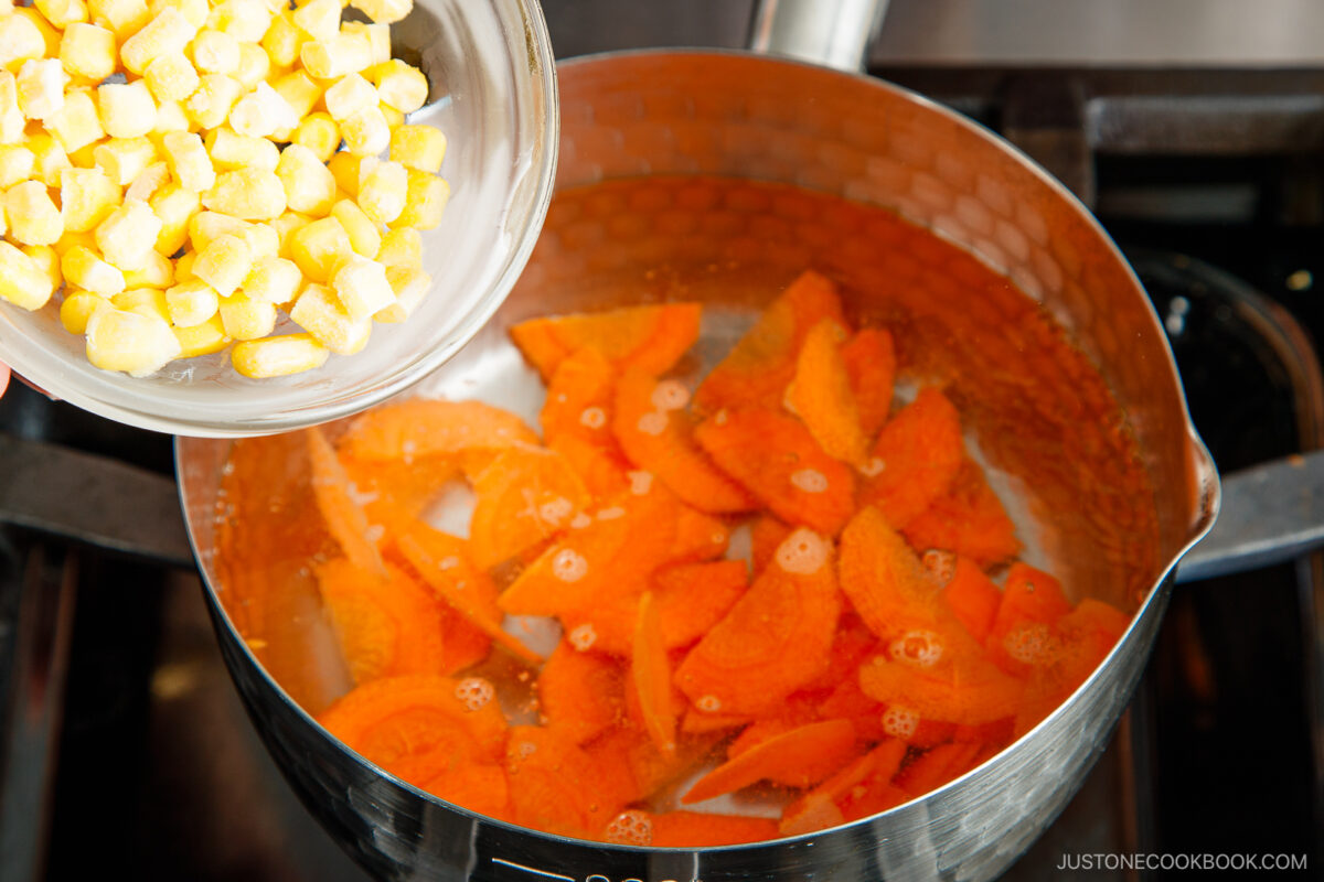 A bowl of frozen corn is being poured into a pot of boiling water with sliced carrots on a stovetop, similar to prepping veggies for japanese potato salad.