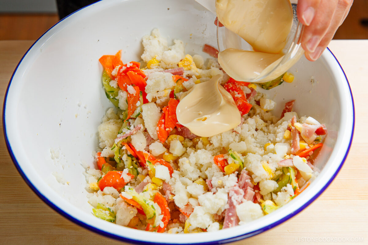 A hand pours creamy dressing from a small bowl over a colorful japanese potato salad filled with cauliflower, carrots, ham, corn, and lettuce in a large white mixing bowl.