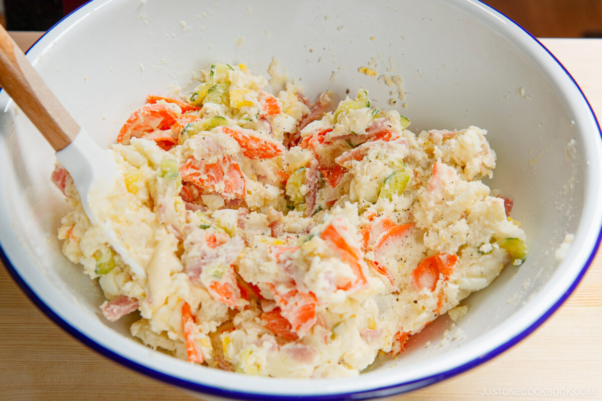A large white bowl filled with Japanese potato salad, featuring chunks of potatoes, carrots, cucumber, and bits of ham, being mixed with a white spatula on a wooden surface.