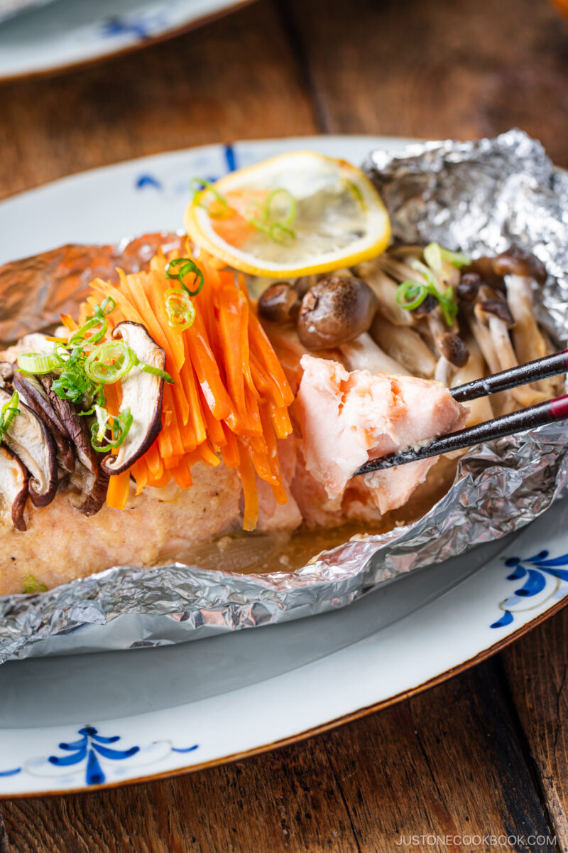 A piece of miso salmon in foil, topped with sliced carrots, mushrooms, and green onions, sits on a plate. Chopsticks hold a bite of salmon, while a lemon slice adds the finishing touch to this flavorful dish.