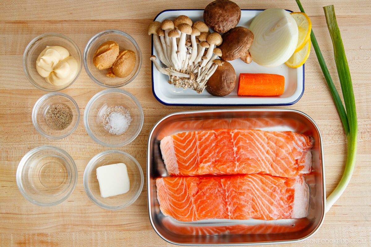 Two raw salmon fillets in a tray, ready for miso salmon in foil, are surrounded by bowls of seasonings, butter, and mayonnaise, plus mushrooms, an onion, carrot, and scallion on a tray—all arranged on a wooden surface.