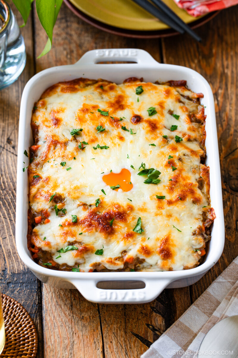 A baked casserole dish filled with cheesy, golden-brown lasagna inspired by Yaki Keema Curry, topped with melted cheese and sprinkled with chopped fresh parsley, set on a rustic wooden table.