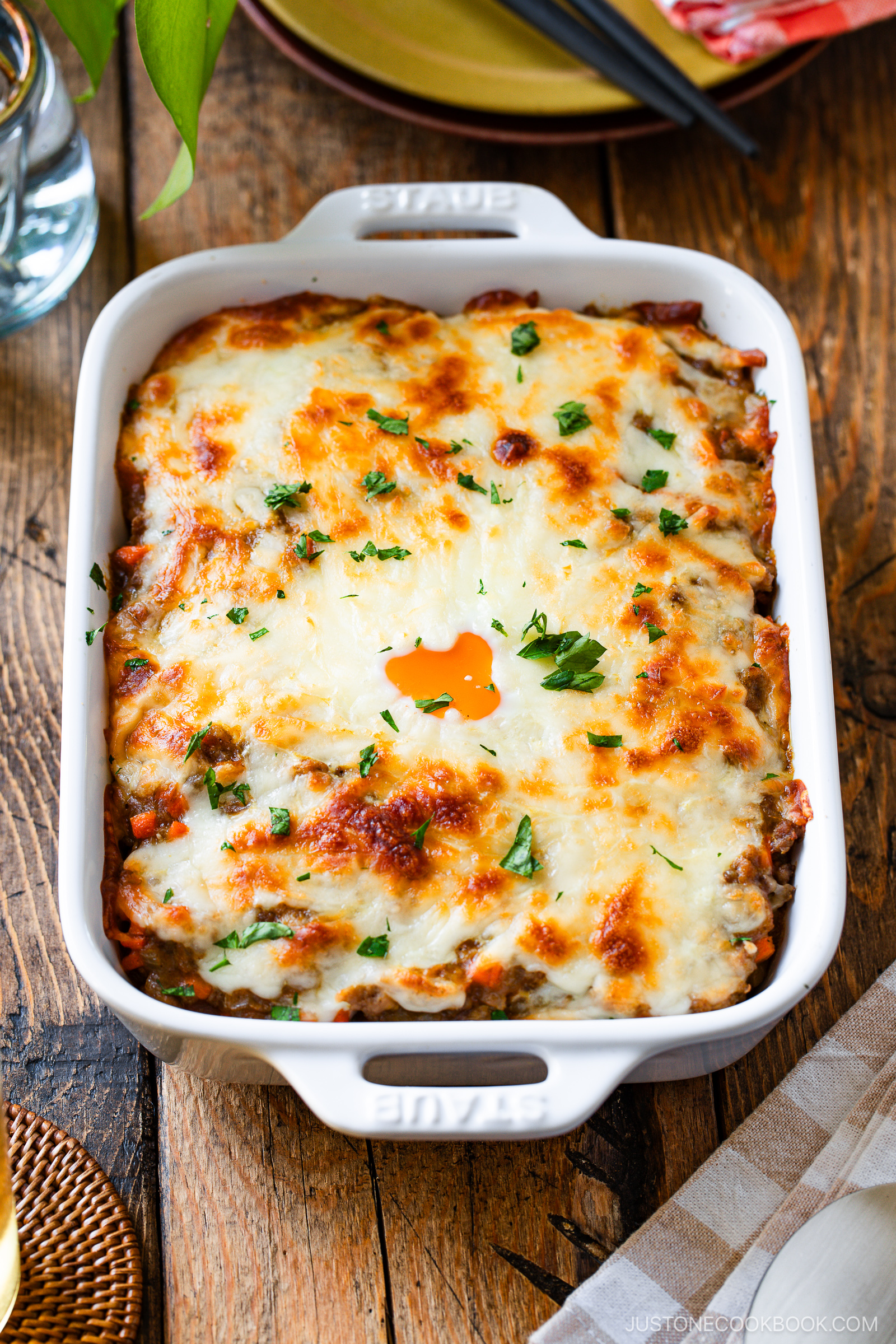 A baked casserole dish filled with cheesy, golden-brown lasagna inspired by Yaki Keema Curry, topped with melted cheese and sprinkled with chopped fresh parsley, set on a rustic wooden table.