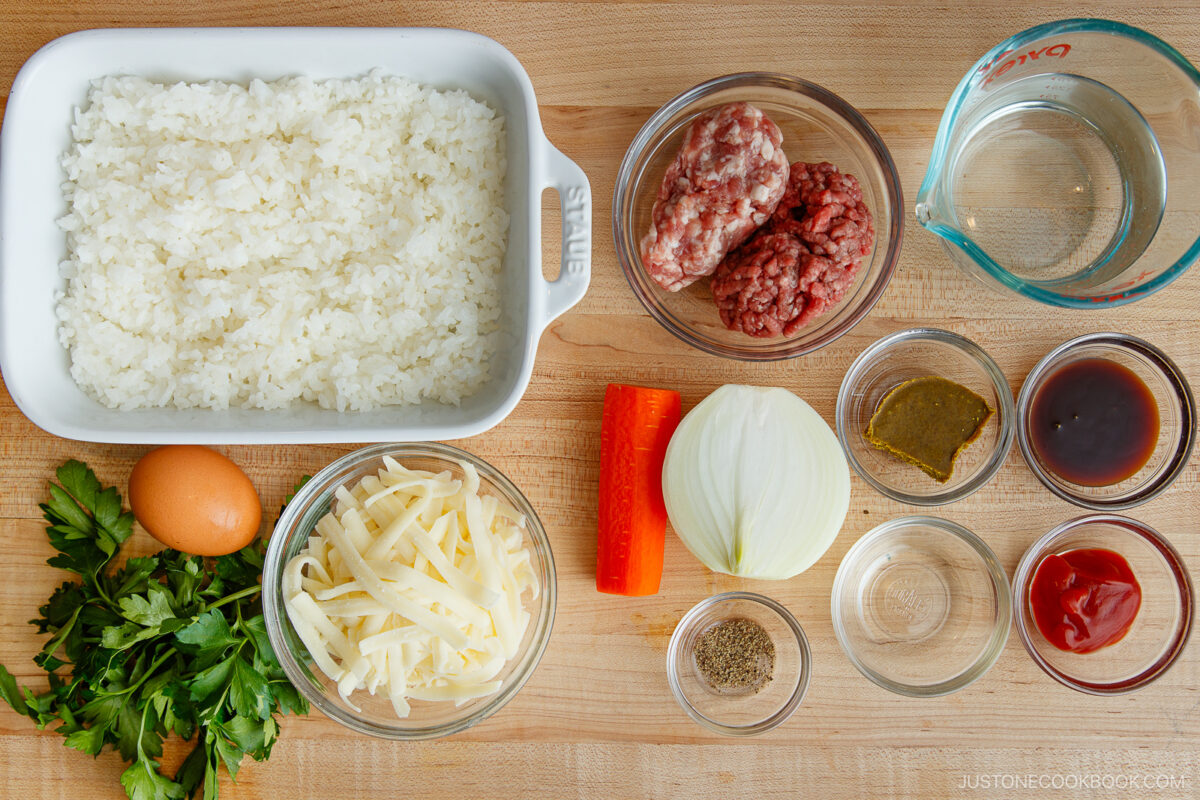 Top-down view of Yaki Keema Curry ingredients on a wooden surface: cooked rice, ground beef and pork, water, half an onion, sliced red bell pepper, egg, parsley, shredded cheese, pepper, bouillon, ketchup, soy sauce, and Worcestershire sauce.