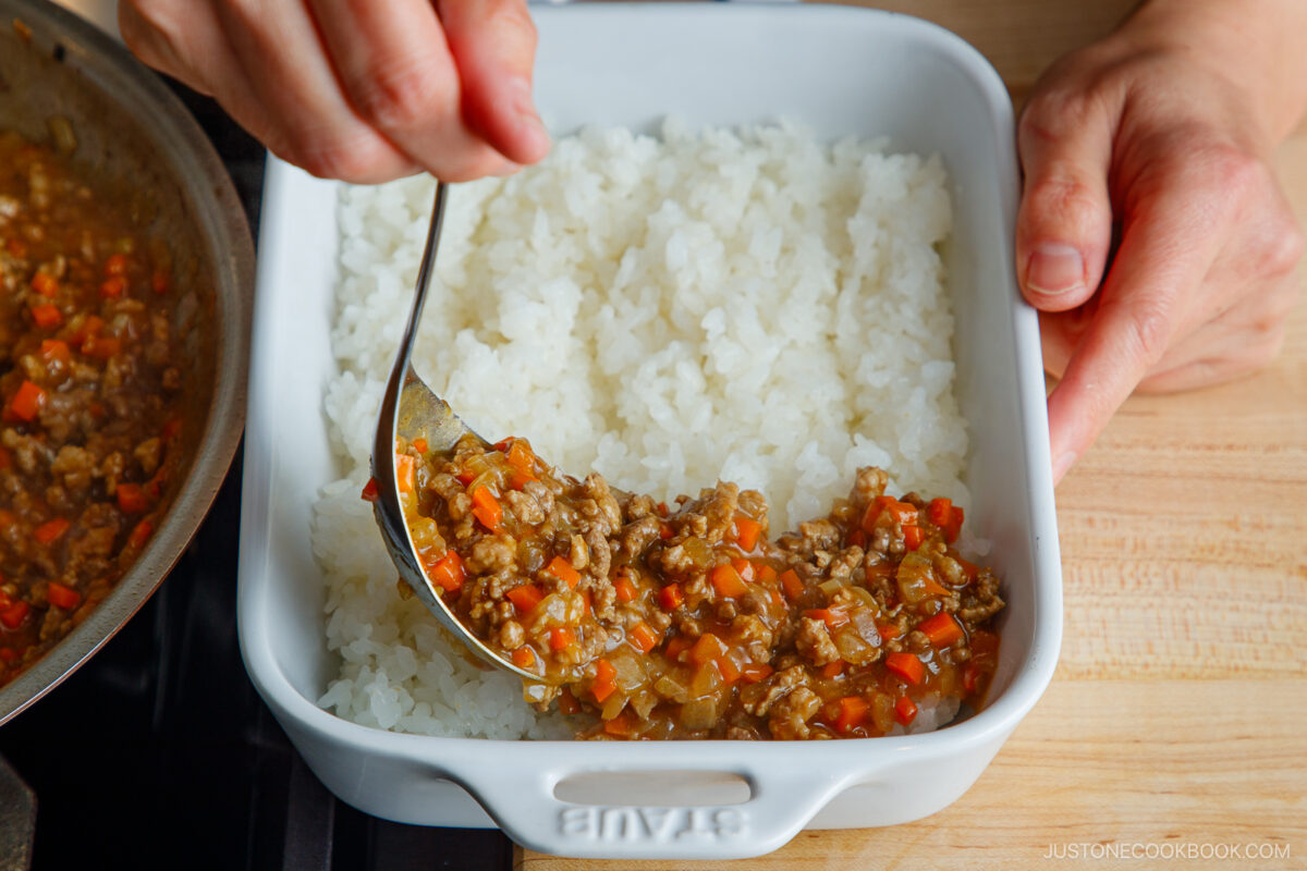 A person spoons a Yaki Keema Curry mixture of ground meat, diced carrots, and onions onto a layer of white rice in a square white baking dish on a wooden surface.