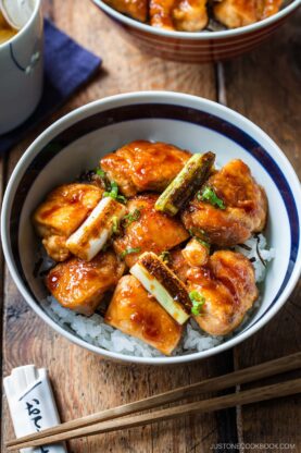 A bowl of steamed white rice topped with glazed, golden-brown yakitori don chicken pieces and grilled green onions, served with chopsticks on a wooden table.