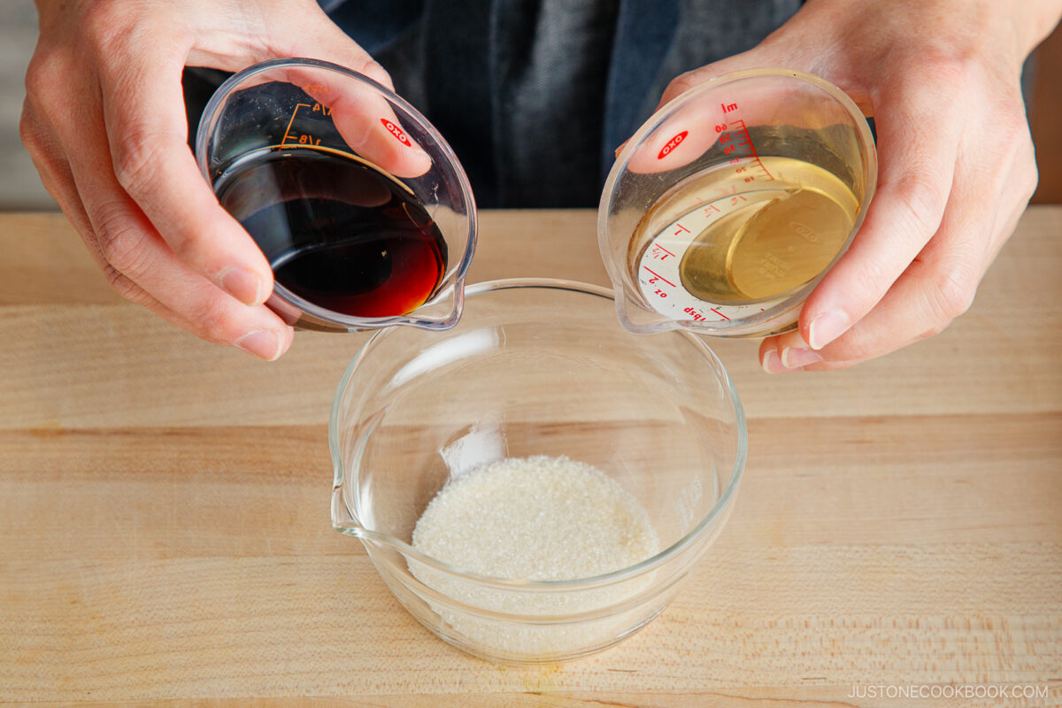 A person holds two measuring cups, one with a dark liquid and one with a light brown liquid, above a glass bowl of granulated sugar—ingredients often used in making yakitori don sauces—on a wooden surface.