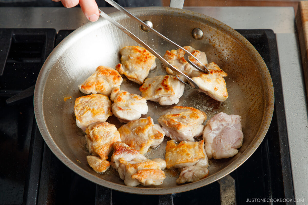 A hand uses tongs to cook and turn pieces of chicken in a stainless steel skillet on a stovetop, preparing savory yakitori don. The chicken pieces are browned and sizzling.