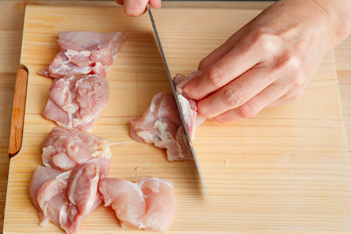 A person slicing raw chicken thigh pieces on a wooden cutting board with a knife, preparing ingredients for yakitori don.