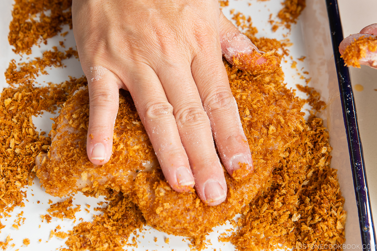 A hand presses a piece of raw chicken into a coating of orange bread crumbs on a white surface, preparing baked chicken katsu for cooking.