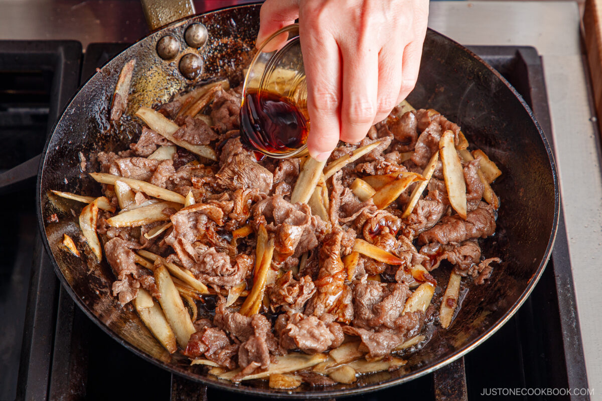 A hand pours dark sauce from a small glass bowl over thinly sliced beef and burdock root for a flavorful Beef and Gobo Stir Fry cooking in a skillet on the stovetop.