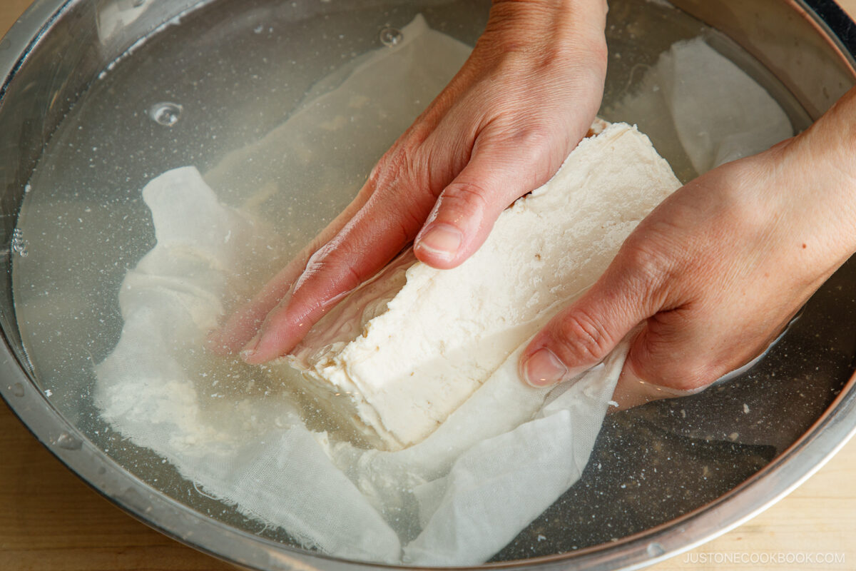 Two hands gently holding and rinsing a block of homemade tofu under water in a large metal bowl, with a white cloth partially wrapped around the tofu.