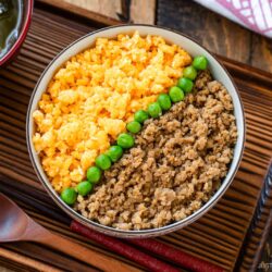 A bowl of soboro don (ground chicken bowl) with scrambled eggs and green peas arranged in neat sections, served on a wooden tray with a wooden spoon and a bowl of miso soup on the side.