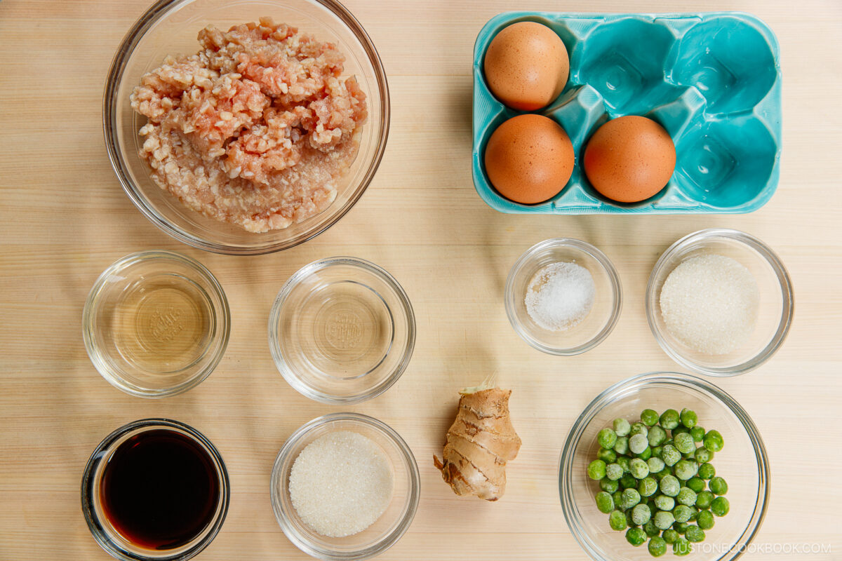A top-down view of ingredients for soboro don (ground chicken bowl) on a wooden surface: ground meat, three brown eggs, soy sauce, rice vinegar, sugar, salt, frozen peas, ginger root, and water in glass bowls.