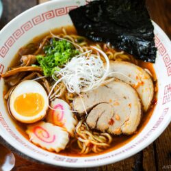 A bowl of spicy shoyu ramen topped with sliced pork, half a soft-boiled egg, green onions, seaweed, bamboo shoots, fish cake slices, and white shredded onions, served in a patterned bowl on a wooden table.