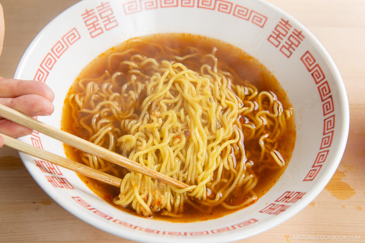 A hand holding chopsticks lifts yellow ramen noodles from a bowl of spicy shoyu ramen, set in a white bowl with a red geometric pattern on the rim.