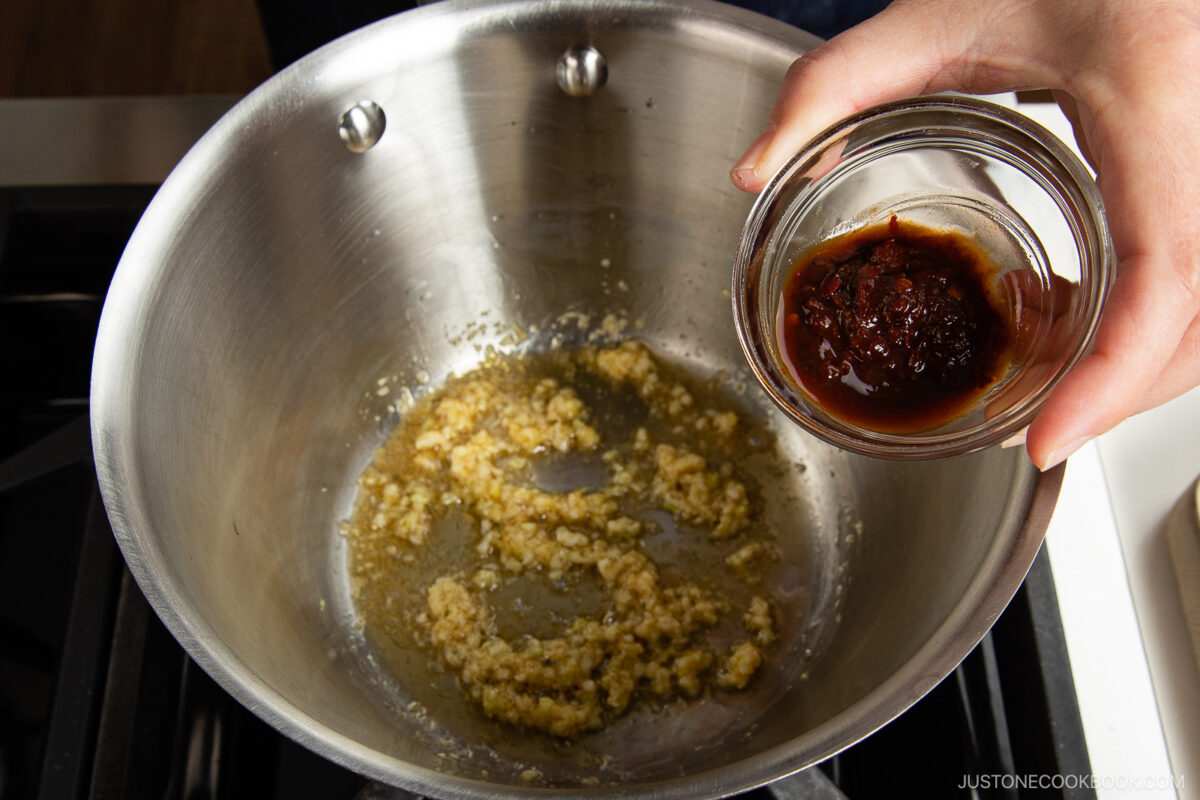 A hand holds a small glass bowl of chili paste above a pot with sautéing minced garlic in oil on a stovetop, preparing the base for spicy shoyu ramen.