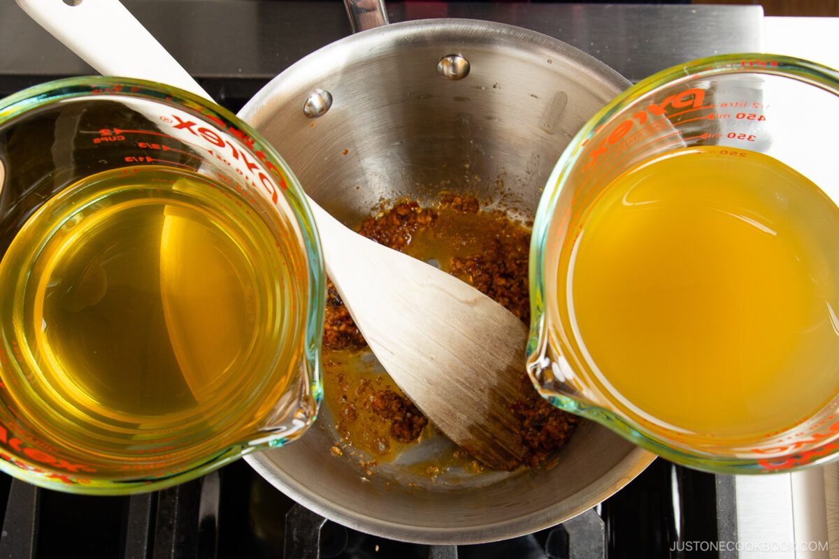 Two glass measuring cups filled with yellow liquid are positioned over a pot of browned spices, being stirred with a wooden spatula on the stovetop—perfect for preparing spicy shoyu ramen.