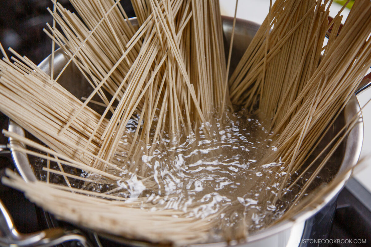 Uncooked spaghetti noodles partially submerged and fanned out in a pot of boiling water on a stovetop, ready to be served alongside zaru soba for a delightful noodle tasting experience.