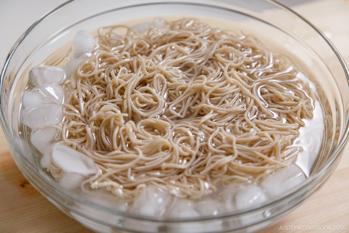 A glass bowl filled with zaru soba soaking in cold water and ice cubes rests on a wooden surface, ready to serve as refreshing cold soba noodles.