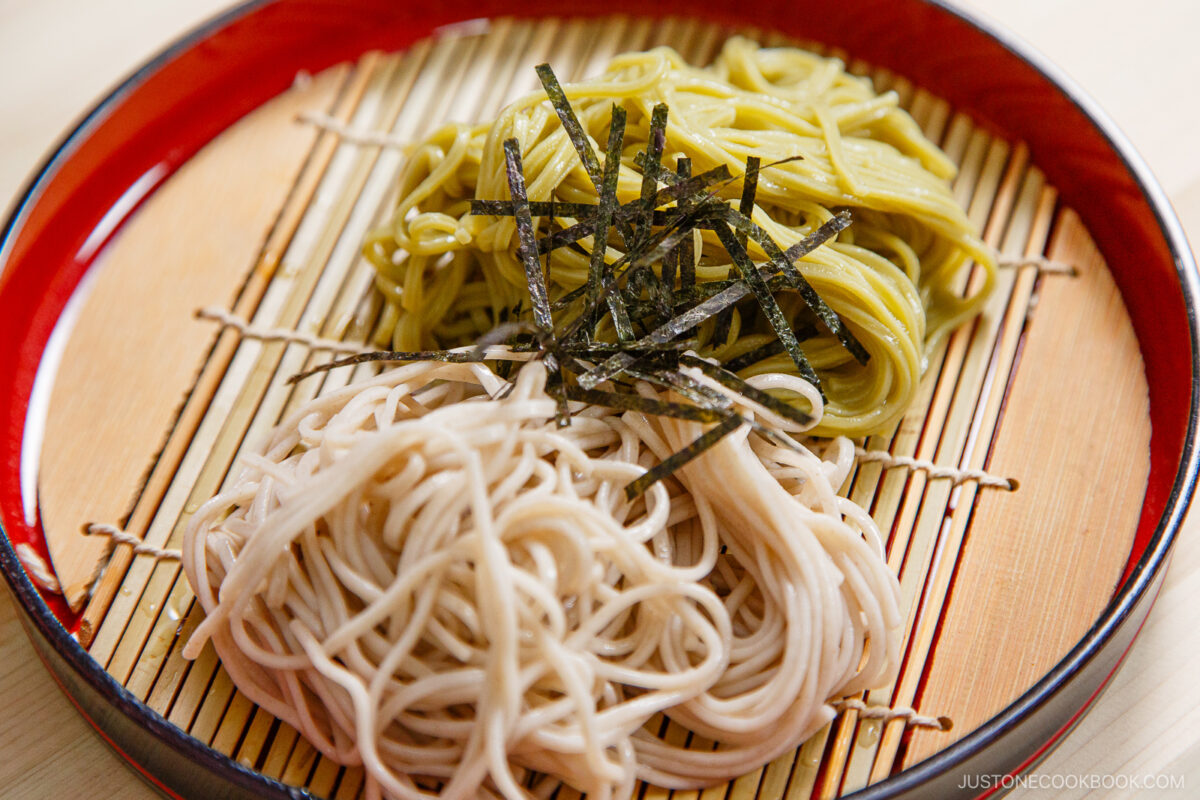 A plate of chilled green tea soba noodles and regular zaru soba, topped with thin strips of nori seaweed, served on a bamboo mat inside a red and black dish.