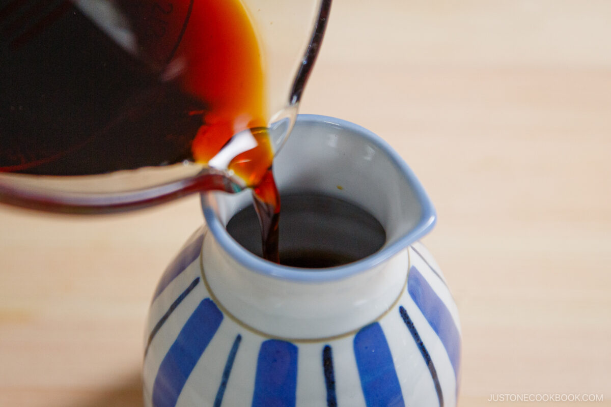 A close-up of soy sauce being poured from a glass container into a small, blue and white striped ceramic pitcher, ready to accompany zaru soba (cold soba noodles), on a light wooden surface.