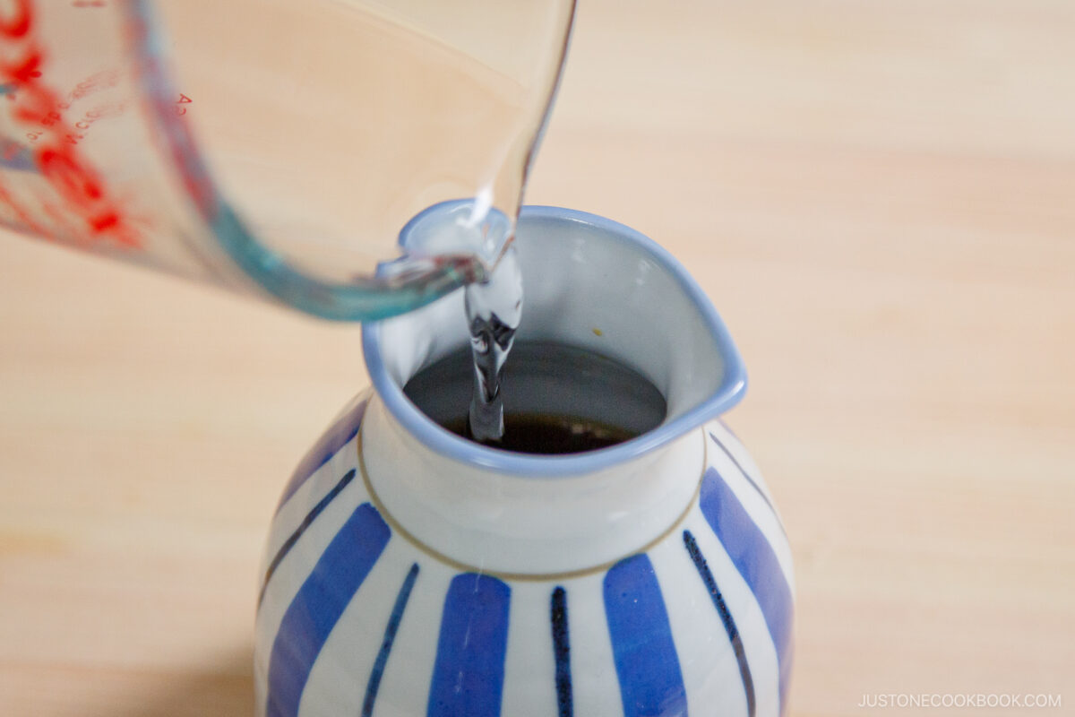 A glass measuring cup is pouring liquid into a small ceramic jug with blue and white stripes on a wooden surface, perfect for serving dipping sauce for zaru soba (cold soba noodles).