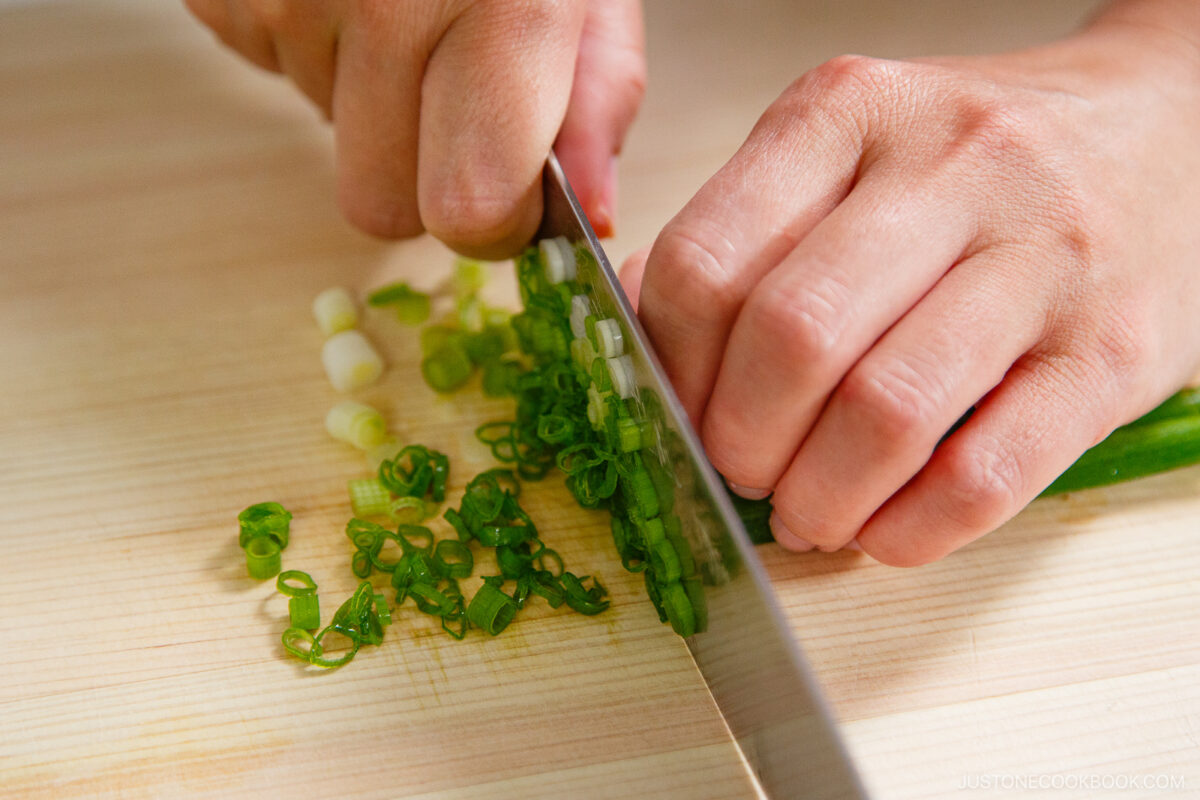 A close-up of hands slicing green onions with a knife on a wooden cutting board, preparing fresh toppings for zaru soba (cold soba noodles).
