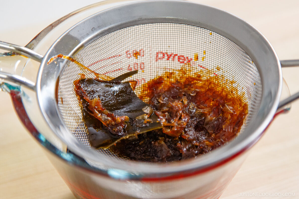A metal strainer sits over a glass measuring cup, holding strained bits of seaweed and bonito flakes used to make Japanese dashi broth—the perfect base for zaru soba (cold soba noodles). The liquid has passed through, leaving the solids behind.