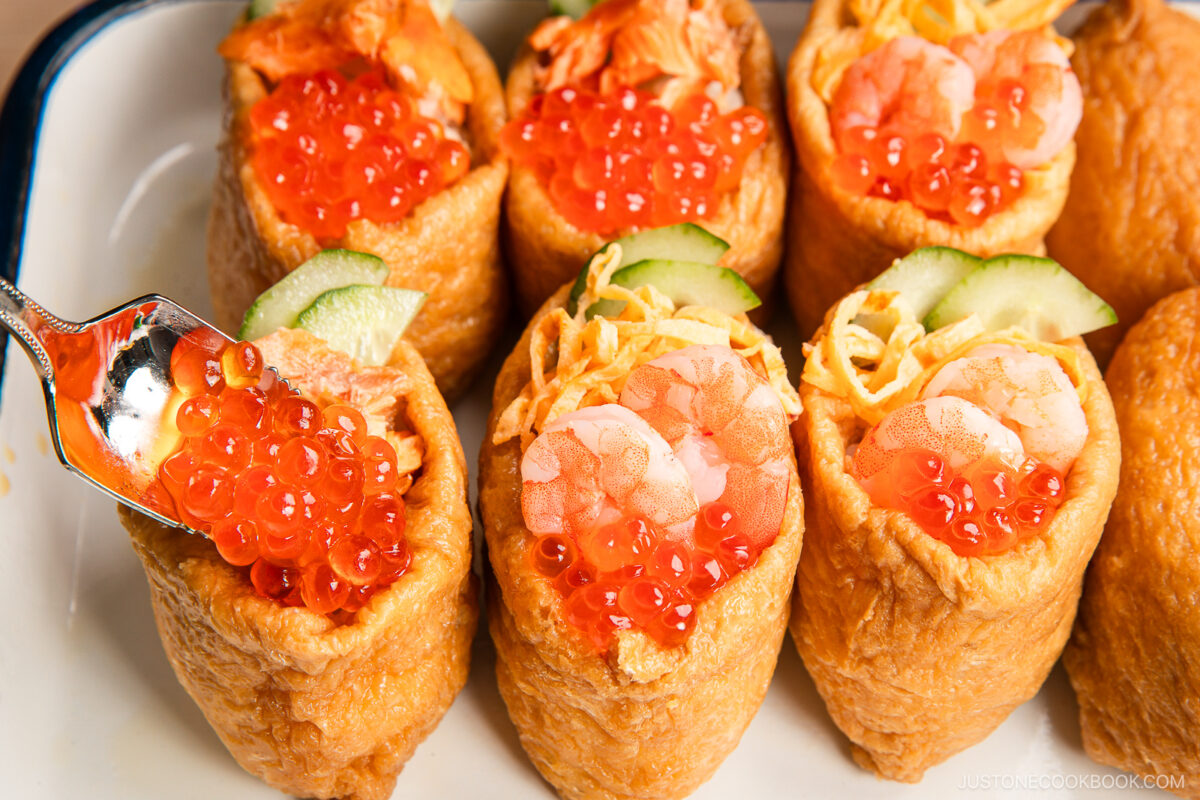 A close-up of inari sushi pouches filled with rice, shrimp, shredded egg, cucumber slices, and topped with bright orange salmon roe, as a spoon adds even more roe to the savory inari sushi creation.