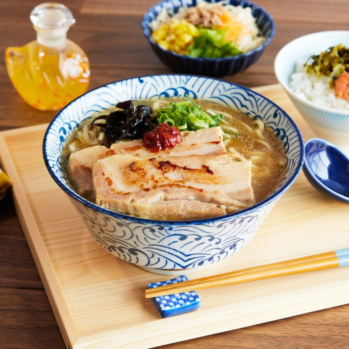 A bowl of ramen with beautiful plating, topped with sliced pork, green onions, mushrooms, and a dollop of chili paste, served on a tray with rice dishes and a glass jar of oil. Chopsticks and spoon rest nearby.