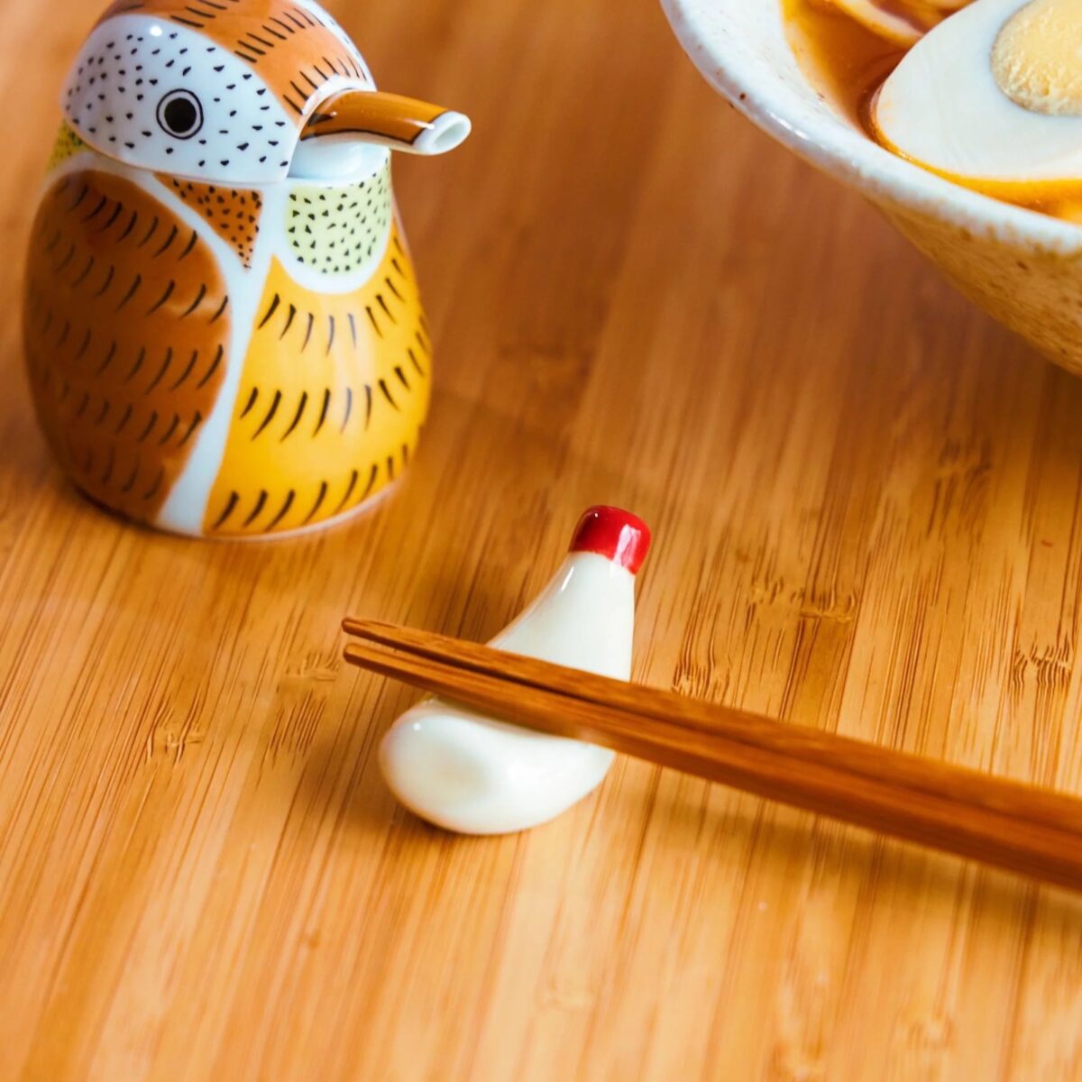 A pair of wooden chopsticks rests on a small ceramic chopstick holder shaped like a bird, next to a bird-shaped soy sauce dispenser and a bowl of ramen with an egg, showcasing beautiful plating on a wooden surface.
