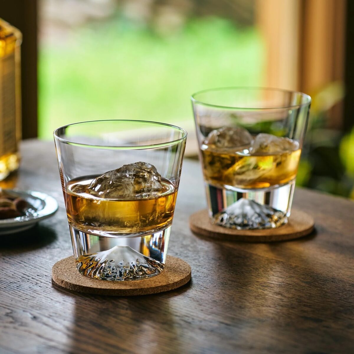 Two glasses of whiskey with ice rest on cork coasters atop a wooden table, their beautiful plating complemented by a blurred green outdoor background visible through the window.