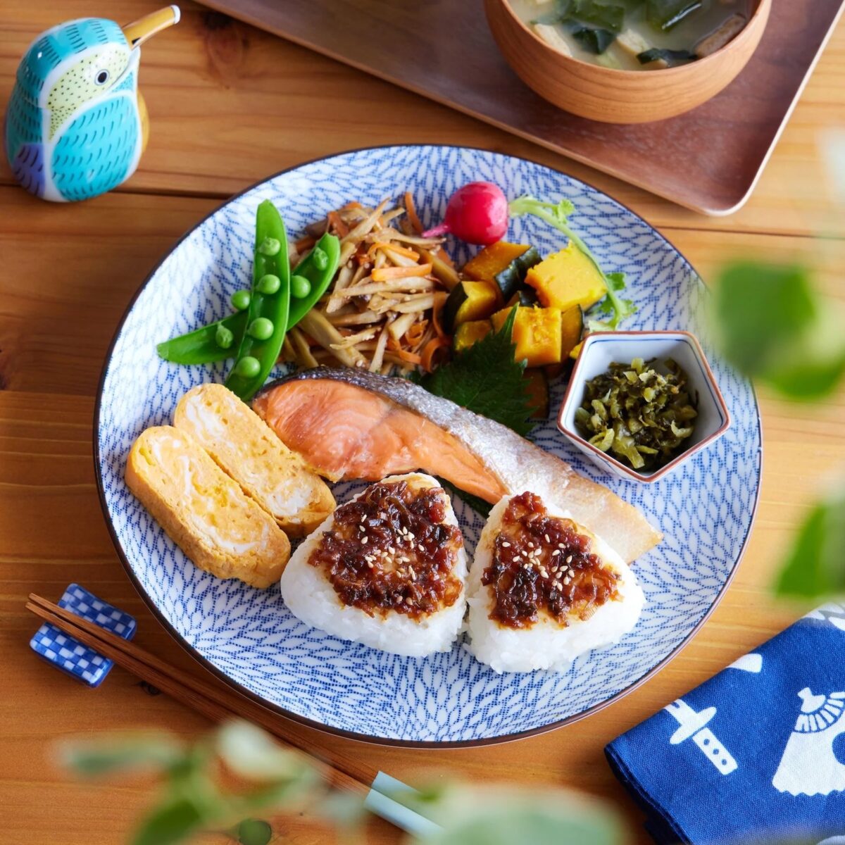 A Japanese breakfast plate with beautiful plating: grilled salmon, tamagoyaki, rice balls with sauce, snow peas, simmered vegetables, pickles, and miso soup arranged neatly on a blue-patterned dish atop a wooden table.
