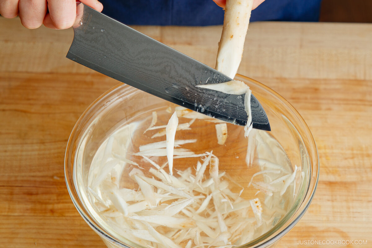 A person does prep work, slicing burdock root into thin strips over a bowl of water with a knife, on a wooden surface.