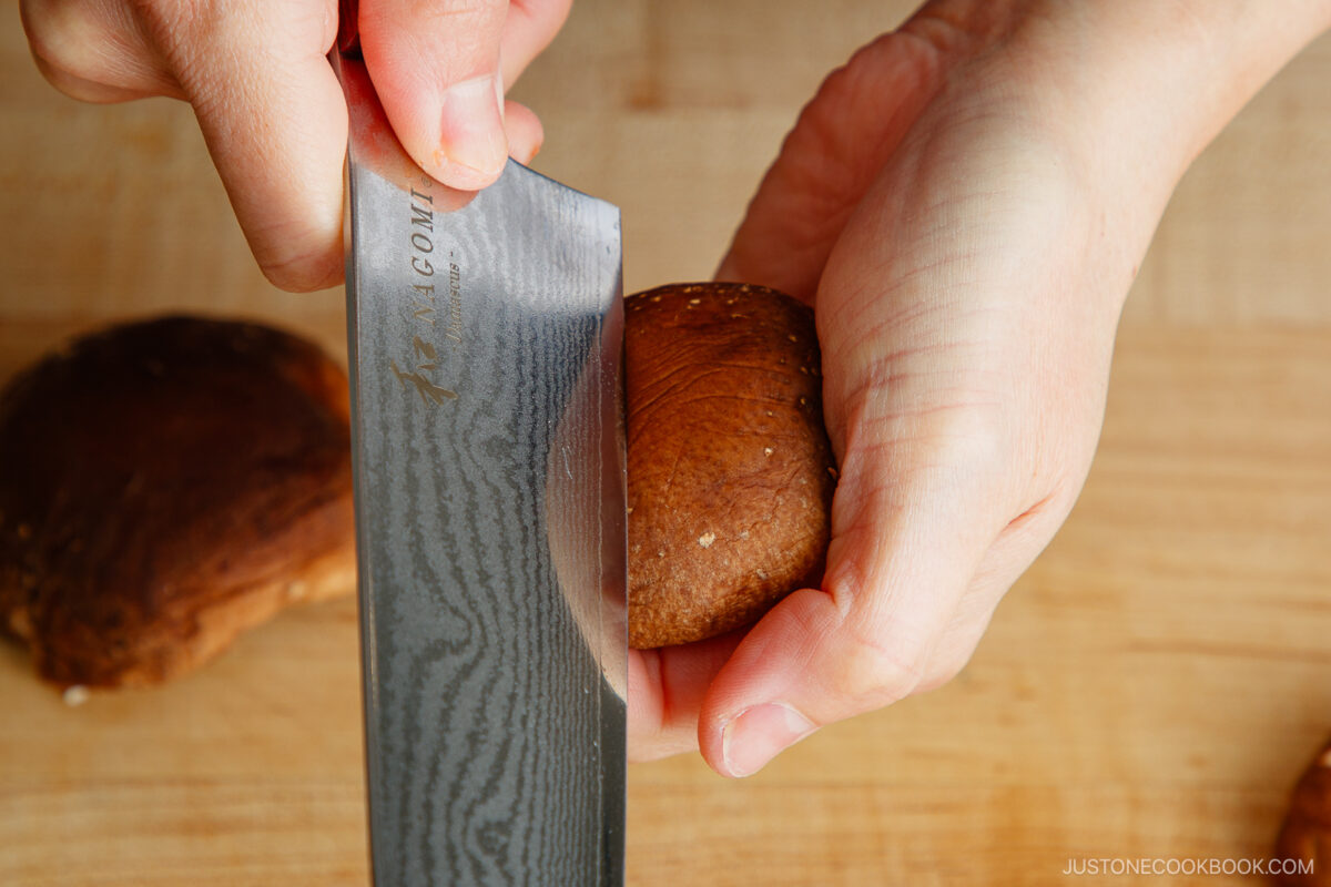 A close-up of hands using a knife to slice the stem off a brown mushroom on a wooden cutting board, showing part of the process for how to make a flower pattern on shiitake mushroom caps.