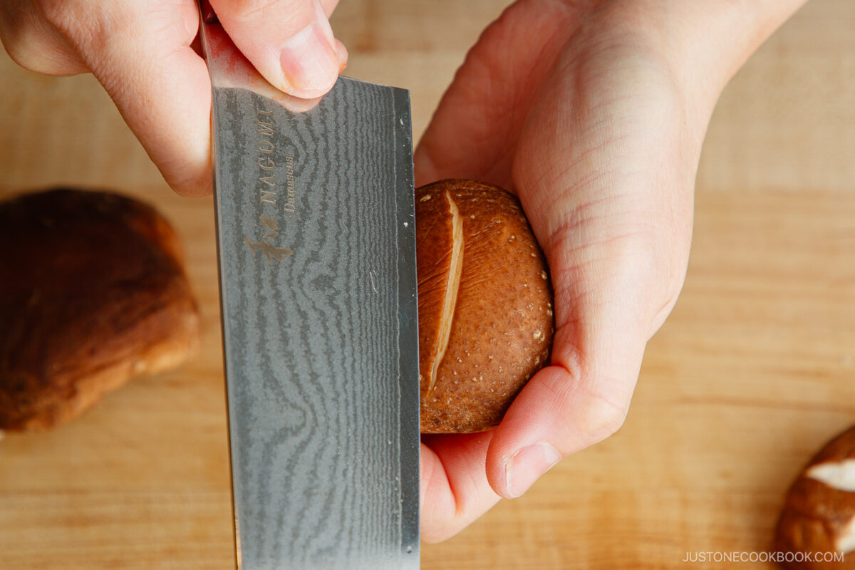 A person demonstrates how to make a flower pattern on shiitake mushroom caps, using a sharp knife to carefully score the top while holding it steady on a wooden cutting board.