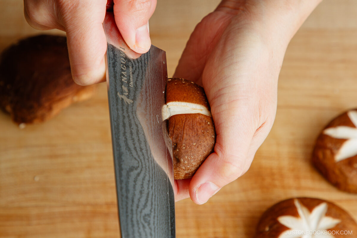 Close-up of hands using a knife to score an X-shaped cut on a brown mushroom cap, demonstrating how to make a flower pattern on shiitake mushroom caps. Other mushrooms with similar decorative cuts are in the background.