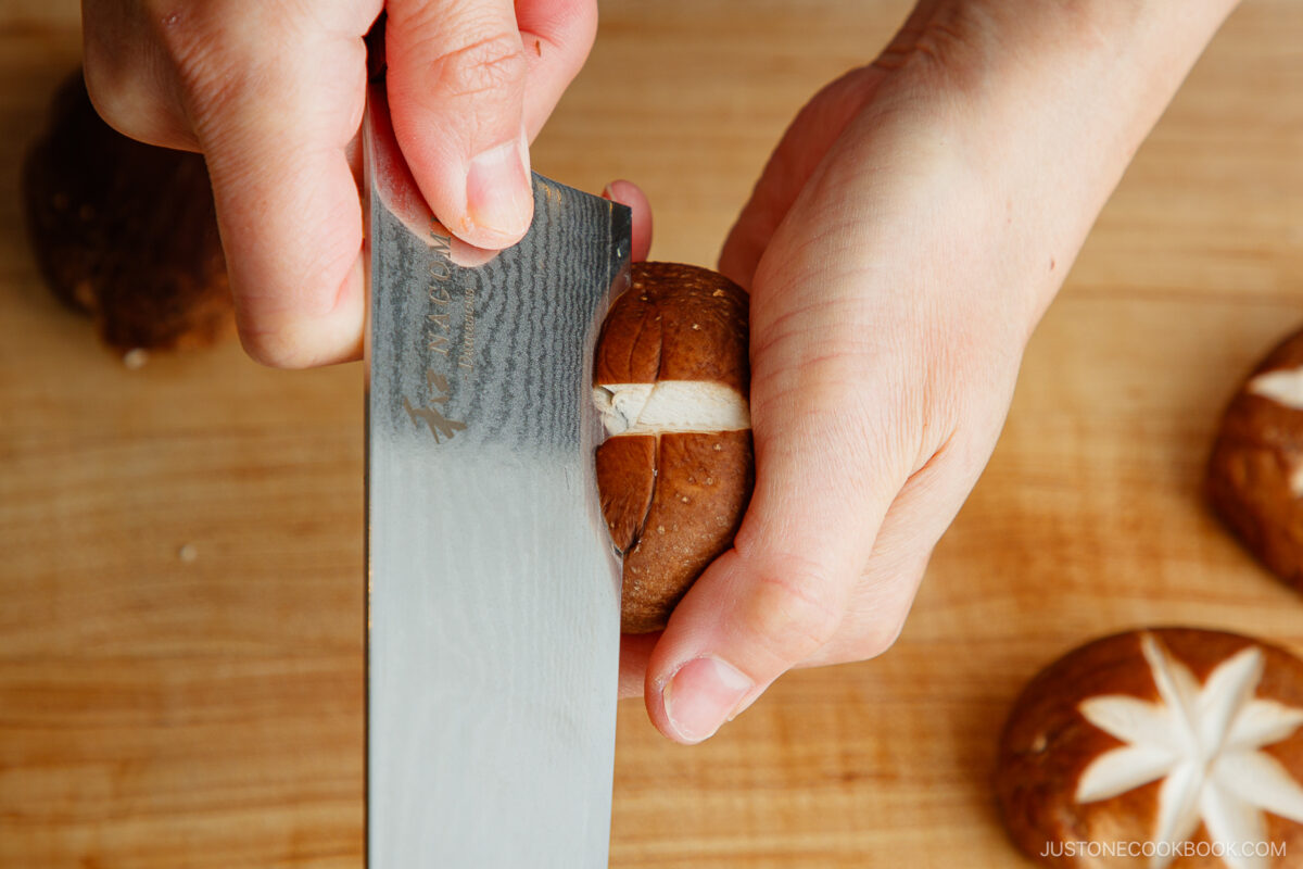 A person demonstrates how to make a flower pattern on shiitake mushroom caps by scoring an X shape with a knife over a wooden cutting board; other mushrooms are nearby.