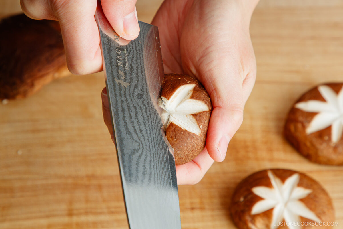 A hand uses a sharp knife to carve a decorative star, demonstrating how to make a flower pattern on shiitake mushroom caps on a wooden cutting board.