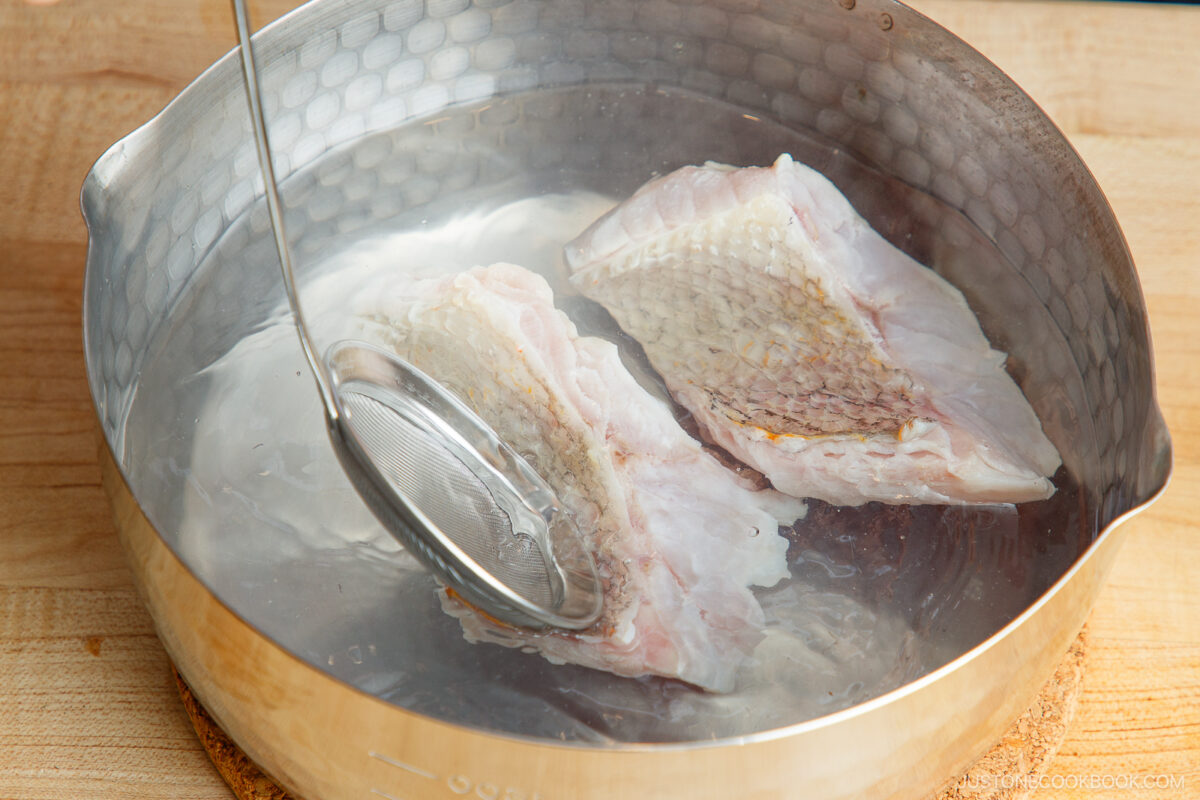 Two raw fish fillets are undergoing prep work as they’re blanched in a pot of hot water, with a metal skimmer partially submerged beside them. The pot rests on a wooden surface.