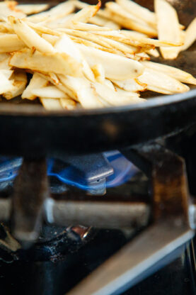 Sliced burdock root being sautéed in a black skillet over a blue gas flame on a stovetop.