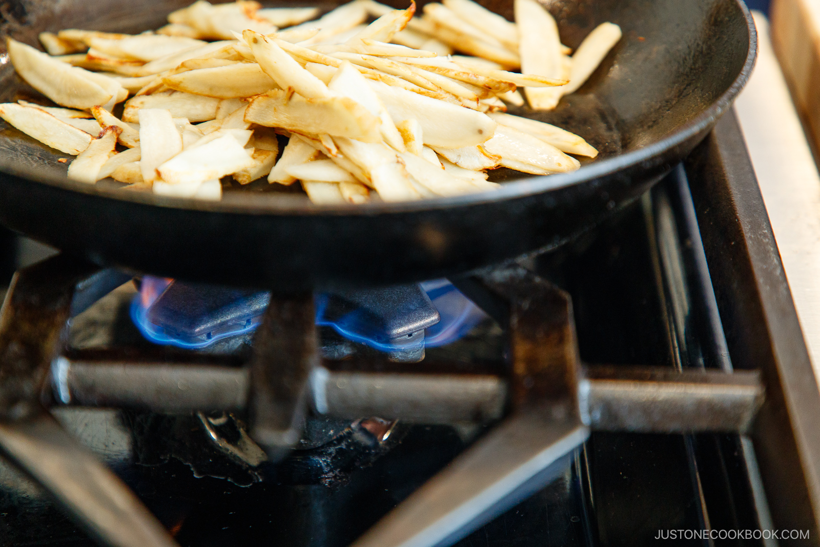 Sliced burdock root being sautéed in a black skillet over a blue gas flame on a stovetop.