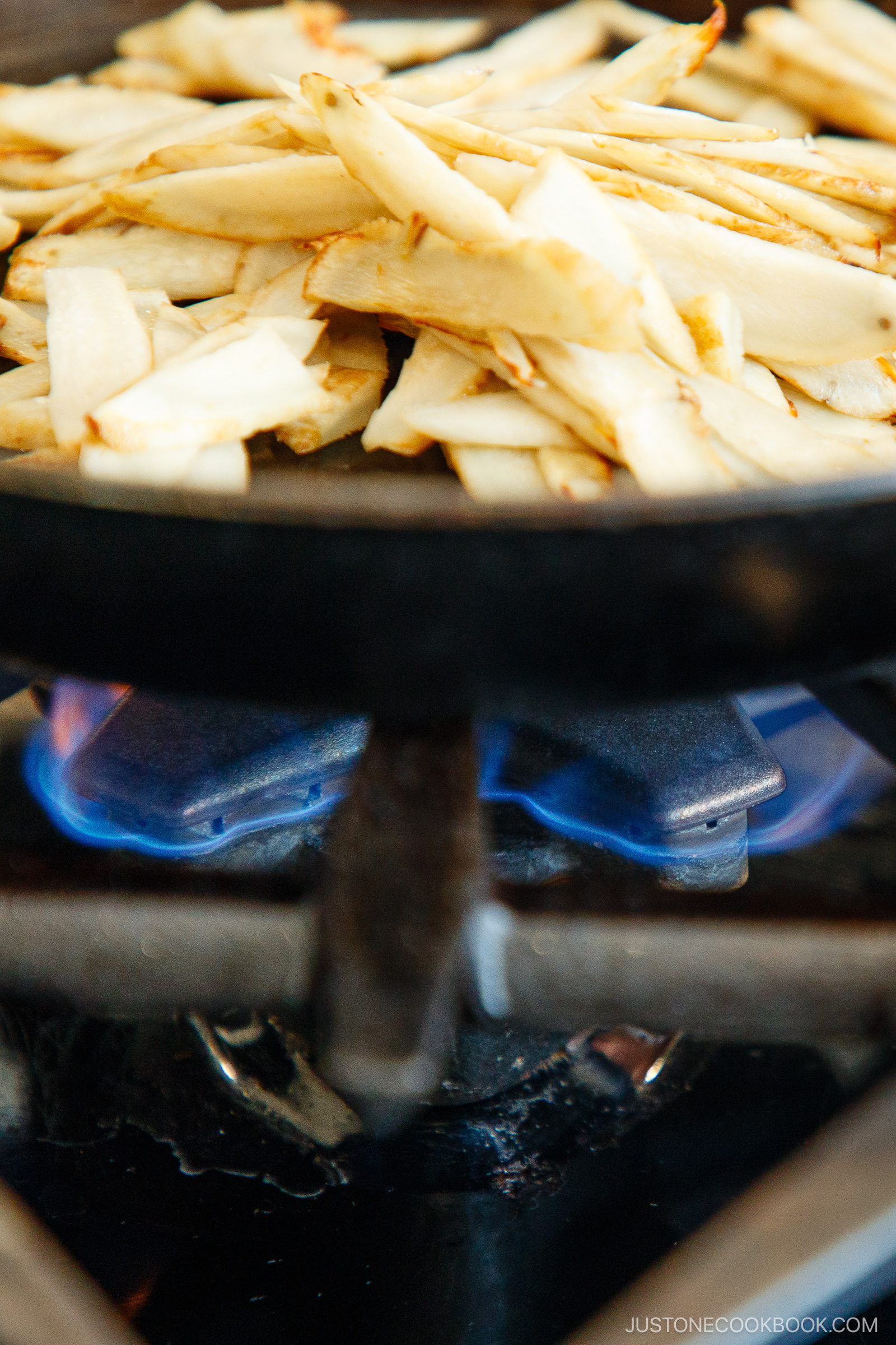 Sliced potatoes cooking in a black skillet over a blue gas flame on a stovetop.