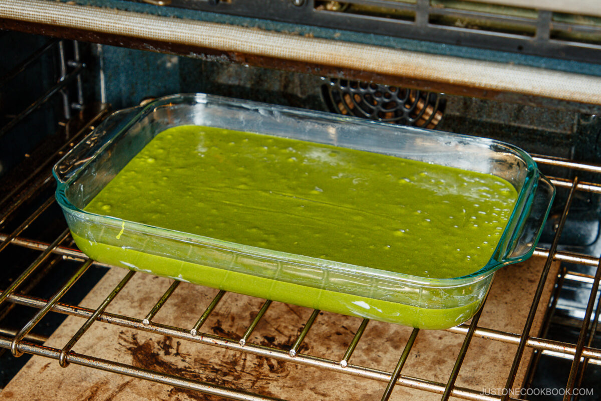 A glass baking dish filled with matcha butter mochi batter sits on the oven rack, ready to be baked. The oven interior is visible, and the vibrant green batter is smooth and evenly spread.