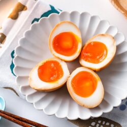 Four ramen eggs, halved to reveal their soft, runny yolks, are arranged on a white scalloped plate set on a patterned cloth. A pair of wooden chopsticks and a blue dish complete this inviting ramen eggs presentation.