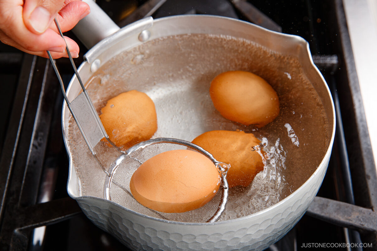 A hand uses a slotted spoon to lift a brown ramen egg from a pot of boiling water on a stove, while three other eggs continue to cook in the pot.