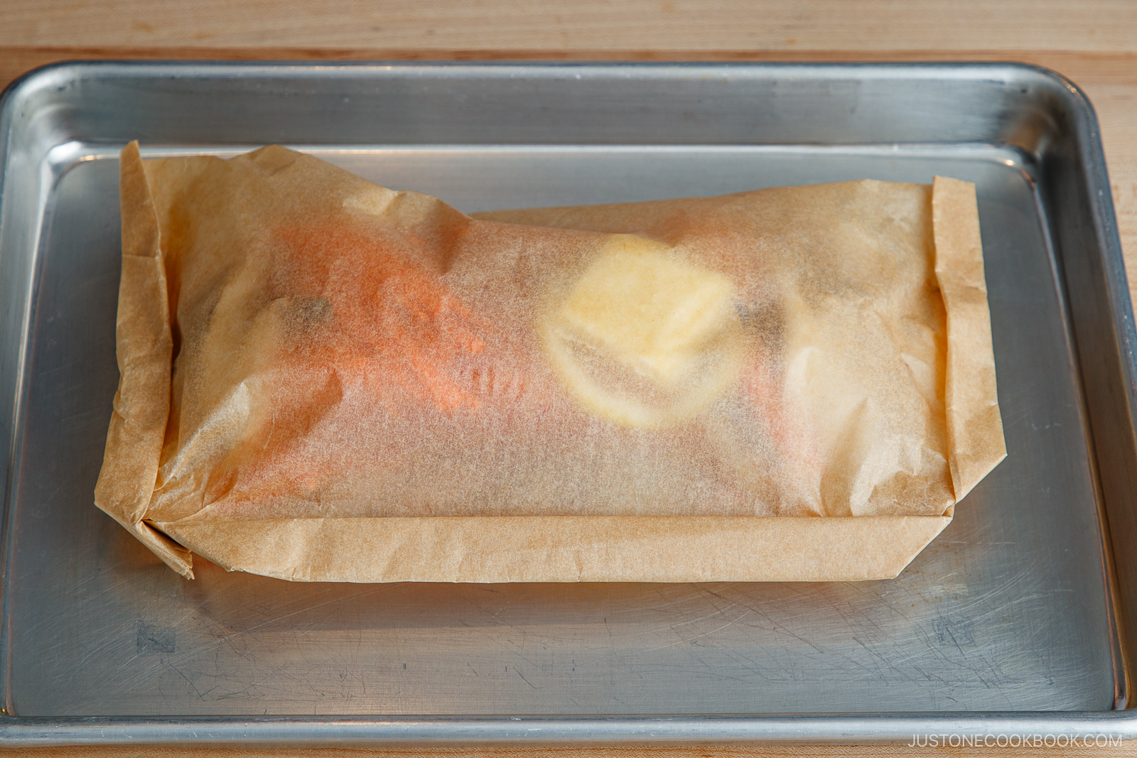 A parchment paper packet with food inside, placed on a metal baking tray—similar to how salmon in foil is cooked. The food contents are faintly visible through the paper.