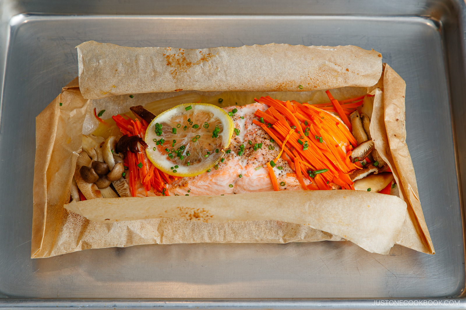 Salmon in foil baked with julienned carrots, mushrooms, and a lemon slice, then garnished with chopped chives, served on a metal baking tray.